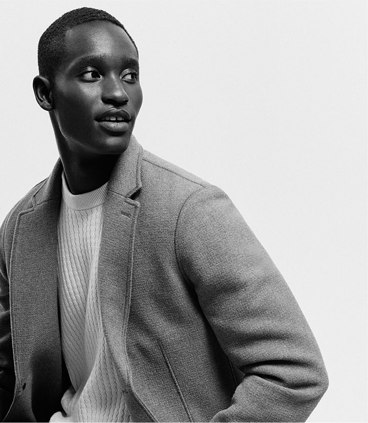 a black and white photo of a man posing wearing a round-neck sweater and a blazer