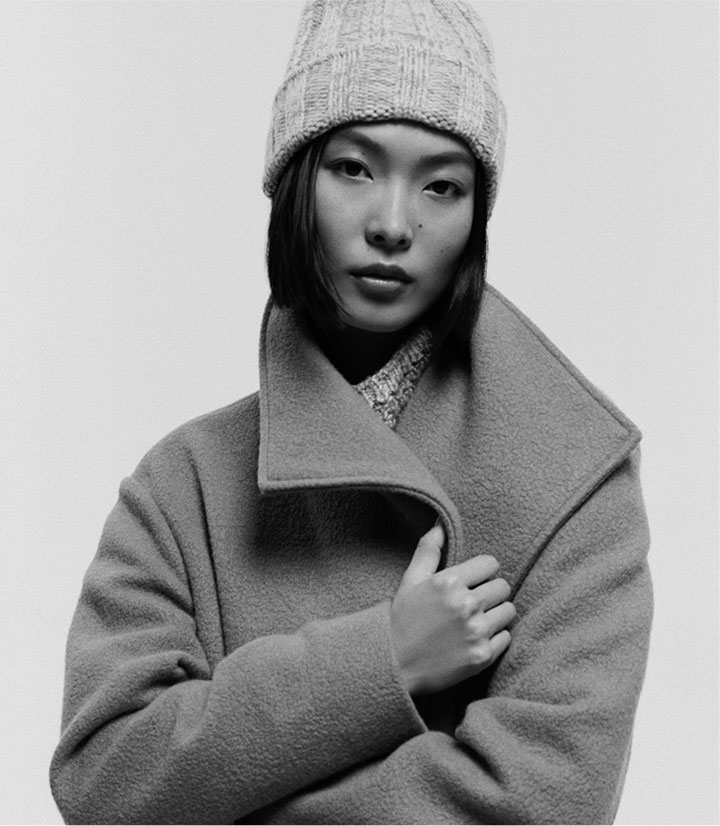 a black and white photo of a woman posing wearing a knitted hat and wool-blend coat